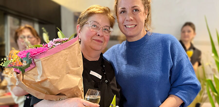Natalie Maurer hält einen großen Blumenstrauß und ein Glas Wein in den Händen und steht neben Jennifer Kürsten