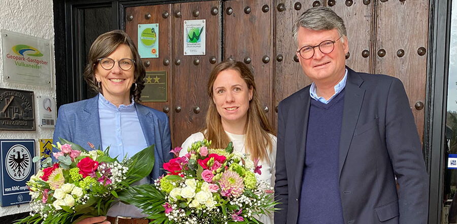Drei Personen mit Blumensträußen vor der hölzernen Eingangstür des Haupthauses