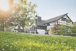 Blick über eine grüne Wiese mit Gänseblümchen hinweg auf das große Haupthaus des Sporthotel-Grafenwald.