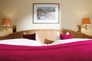 Blick über das Fußende hinweg zum Kopfende des Doppelbetts im Ferienhaus. Links und rechts eine Wandlampe, über dem Kopfteil ein Bild.