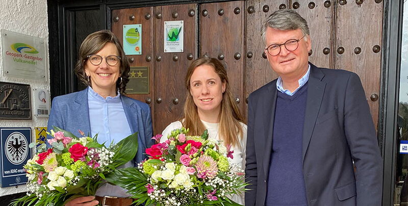 Drei Personen mit Blumensträußen vor der hölzernen Eingangstür des Haupthauses
